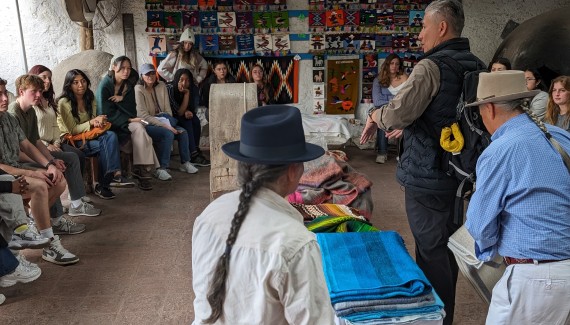 Cornell students sit around a teacher in Ecuador displaying a large tool for weaving. 