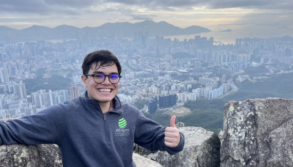 Students smiles and holds a thumbs up above the Hong Kong skyline.
