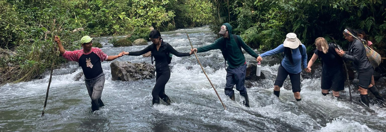 Students in Ecuador are led by a guide across a river. They link arms and cross in a single-file line.