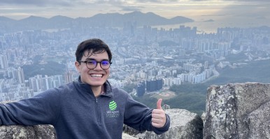 Students smiles and holds a thumbs up above the Hong Kong skyline.