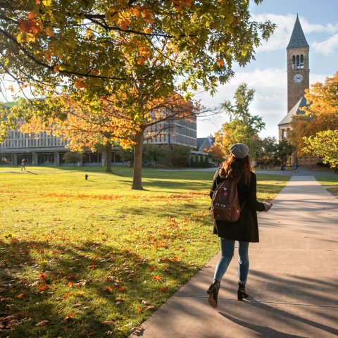 A female student wearing a hat and backpack walks towards McGraw Tower on a sunny autumn day
