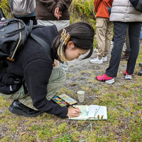 Student squats over a notebook where she paints a watercolor picture of the landscape. 